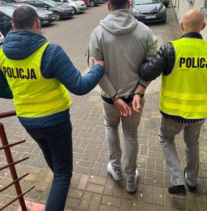 Dwaj funkcjonariusze prowadzą zatrzymanego mężczyznę.