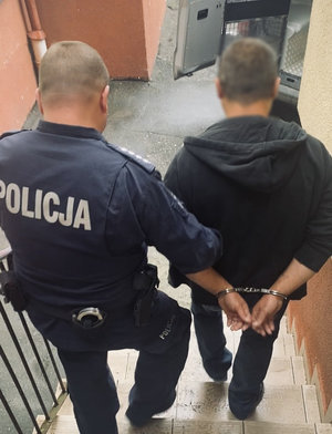 Policjant prowadzi zatrzymanego mężczyznę zakutego w kajdanki.