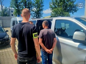 Zatrzymany mężczyzna w kajdankach z policjantami przy radiowozie.