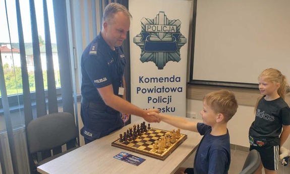Komendant Powiatowy Policji w Lesku młodszy inspektor Grzegorz Koczera podaje rękę chłopcu przed grą w szachy. Między nimi stolik z szachami.