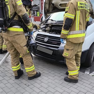 Na zdjęciu widocznych jest dwóch umundurowanych strażaków stojących obok białego busa, który ma podniesiona maskę.