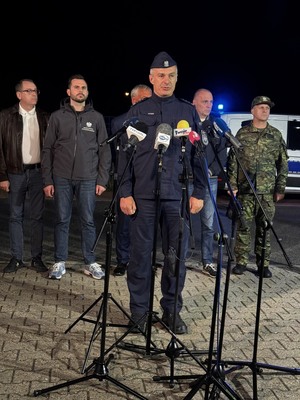 Policjant przed mikrofonami na konferencji prasowe. Za nim widocznych stojących pięciu mężczyzn, biorących również udział w konferencji prasowej.