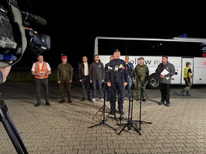 Policjant przed mikrofonami na konferencji prasowe. Za nim widocznych stojących pięciu mężczyzn, biorących również udział w konferencji prasowej.