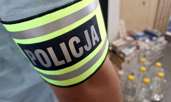 Na pierwszym planie widoczna opaska na ramieniu z napisem Policja. w tle widać zabezpieczone butelki z nielegalnym alkoholem.