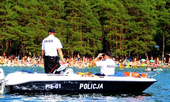 policjanci stoją na łódce i patrolują okolice. Na plaży w tle siedzi dużo ludzi.