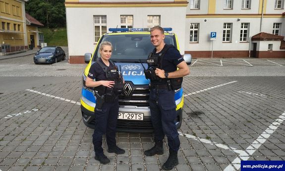 Dwoje umundurowanych policjantów przed radiowozem.