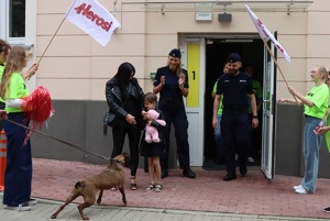 Dziewczynka i kobieta przed wejściem do szpitala, obok stoi policjant i policjantka, po bokach młode kobiety z białymi flagami, na których znajduje się czerwony napis Herosi.