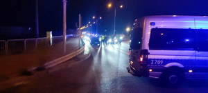 Na ulicy oświetlone pojazdy i policjanci kontrolujący auta. Czynności wykonywane w porze nocnej.