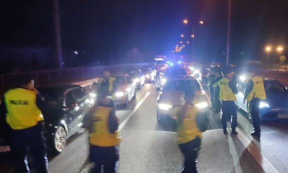 Na ulicy oświetlone pojazdy i policjanci kontrolujący auta. Czynności wykonywane w porze nocnej.