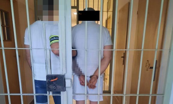 Stojącego tyłem mężczyznę trzyma policjant z opaską na ręce POLICJA. Obaj stoją za kratami.