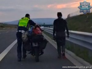 policjant prowadzi motocykl, obok idzie motocyklistka.