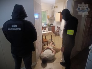 Zatrzymany mężczyzna leży na podłodze zakuty w kajdanki. Obok niego stoi funkcjonariusz służby celno-skarbowei i policjat.