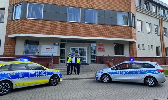 Przed budynkiem komisariatu stoją dwa radiowozy, oraz dwaj policjanci z mężczyzną.
