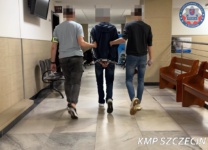 Dwaj funkcjonariusze prowadza zatrzymaną osobę korytarzem.