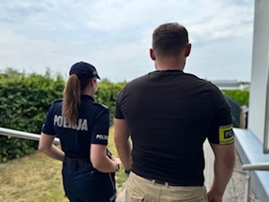 Umundurowana policjantka i ubrany po cywilnemu policjant z opaska na ręce z napisem Policja. Widok z tyłu.