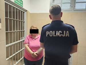 Zatrzymana kobieta i stojący tyłem policjant, u którego widać napis na plecach: Policja.