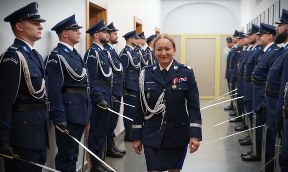 Policjantka idzie środkiem korytarza. Po obu stronach wzdłuż korytarza stoją policjanci z szablami.