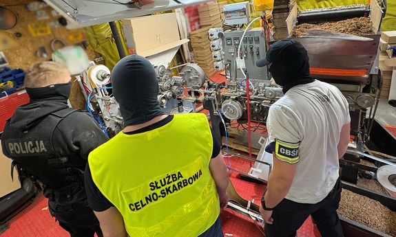 Policjant i funkcjonariusz KAS i CBŚP stoją przed maszyna do produkcji nielegalnych papierosów.