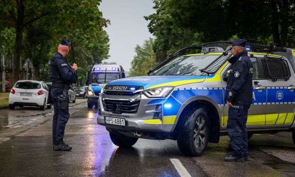 Dwaj policjanci przy radiowozie. na drugim planie widać nadjeżdżający policyjny furgon, a z lewej strony zaparkowane cywilne auto.