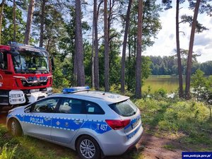 Na pierwszym planie wóz strażacki i policyjny radiowóz. w tle widać zbiornik wodny.