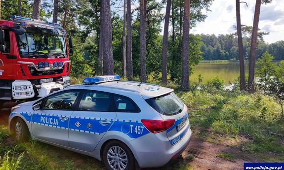 Na pierwszym planie wóz strażacki i policyjny radiowóz. w tle widać zbiornik wodny.