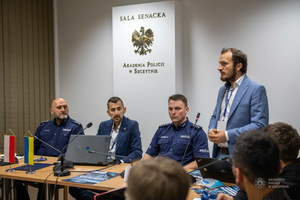 Mężczyzna w garniturze stoi, przy nim siedzą uczestnicy szkolenia.