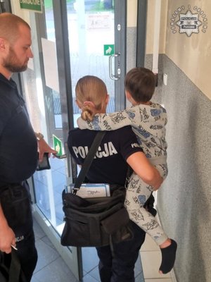 Policjantka trzyma na ręku dziecko. Stojący obok mężczyzna otwiera jej drzwi.