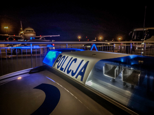 Napis policja na belce na dachu radiowozu. W tle samolot.
