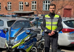 mł. asp. Kamil Styrna, policjant Wydziału Ruchu Drogowego Komendy Miejskiej Policji w Krakowie stoi obok motocykla. W tle radiowozy policyjne.