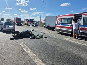 Miejsce wypadku. Na drodze leży motocykl, obok niego stoi karetka pogotowia i samochód osobowy. W głębi wóz strażacki i samochód ciężarowy.