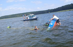 Trzy osoby w wodzie obok wywróconej łodzi. W głębi łódź policyjna.