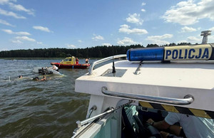 Na pierwszym planie łódź policyjna. W głębi Wywrócona żaglówka i łódź WOPR. Wokół łodzi ludzie w wodzie.