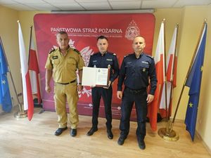 Kłodzki policjant, który podjął ewakuację osób z płonącego lokalu gastronomicznego  trzyma list od Komendanta Powiatowego Państwowej Straży Pożarnej w Kłodzku st. bryg. Rafała Chorzewskiego stojącego obok oraz Komendanta Powiatowego Policji w Kłodzku mł. insp. Tomasza Ociepy.