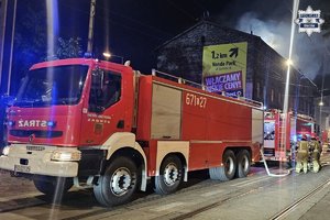 Wozy strażackie i strażacy przy płonącym budynku.