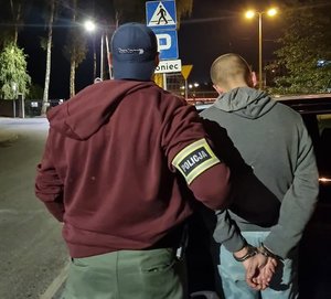 Dwie osoby stojące tyłem. osoba z lewej strony na rękawie posiada opaskę z napisem Policja. Osoba z prawej strony w kajdankach na rękach z tyłu.