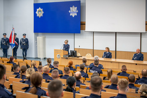 Uroczyste rozpoczęcie szkolenia zawodowego podstawowego. Sala na której widać siedzących słuchaczy. Przodem do nich na podium z lewej strony stoi poczet sztandarowy, przy mównicy widać oficer Policji a za stołem prezydialnym dwoje oficerów Policji.
