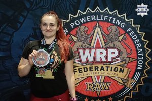 Policjantka w stroju sportowym z medalami na szyi. Jeden z nich prezentuję trzymając w dłoni.
