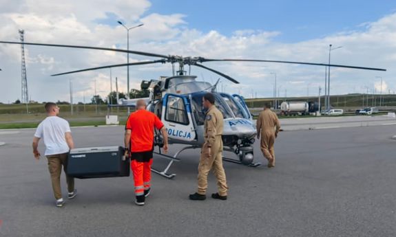 Dwaj mężczyźni niosą skrzynię do śmigłowca. Przy maszynie widać dwóch pilotów.