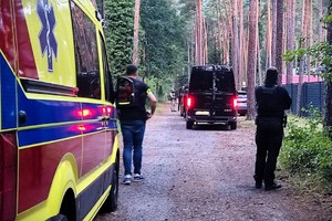Na pierwszym planie widoczna karetka i stojący przy niej mężczyzna, z boku widać policjanta a w oddali policjantów i samochody zaparkowane przed posesją.