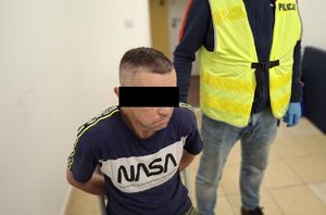 Zatrzymany siedzi na krześle skuty w kajdanki, obok stoi policjant.