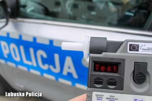 Trzymany w dłoni alkotester. W tle napis policja na radiowozie.