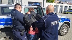 Dwaj funkcjonariusze prowadzą zatrzymanego mężczyznę zakutego w kajdanki.