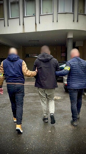Dwaj funkcjonariusze prowadzą zatrzymanego w kajdankach w stronę budynku.
