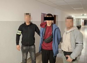 Zatrzymany mężczyzna zakuty w kajdanki z dwoma funkcjonariuszami na korytarzu.