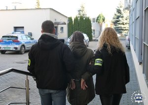 Policjant i policjantka prowadzą zatrzymaną kobietę zakutą w kajdanki.