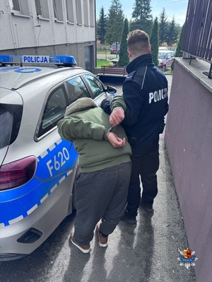 Przed budynkiem placówki oświatowej umundurowany policjant prowadzi do radiowozu zatrzymanego mężczyznę.
