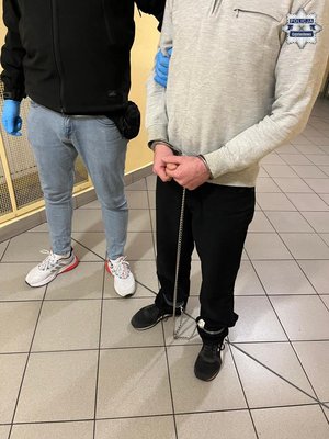Zatrzymany w kajdankach zespolonych trzymany pod rękę przez nieumundurowanego policjanta.