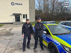 Dwaj policjanci stoją przy radiowozie zaparkowanym przed budynkiem komendy Policji.