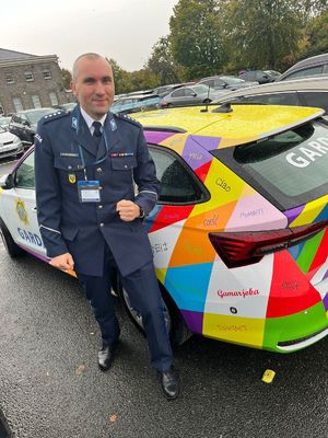 Funkcjonariusz polskiej Policji w umundurowaniu służbowym stoi obok radiowozu policji irlandzkiej. Samochód ozdobiony jest kolorową grafiką z napisami „dzień dobry” w różnych językach, symbolizującą różnorodność kulturową i otwartość irlandzkiej Policji.