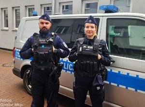 Policjant i policjantka stoją przed radiowozem.